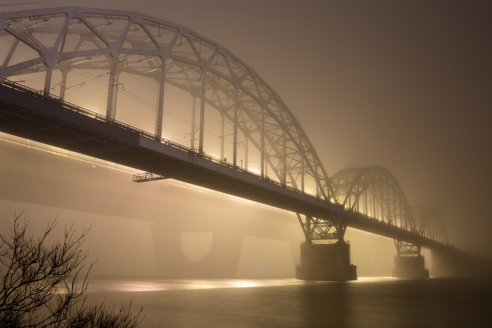 A mysterious evening fog above the river in big city. Bridge in the mist, cold weather scenery. Soft, blurry, misty look. Colorful, mystic industrial cityscape.